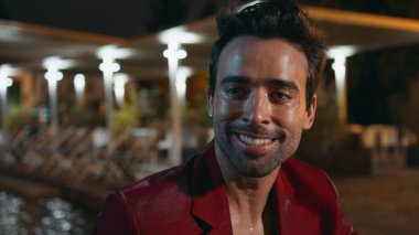 İtalyan adam parti gecesi konuşuyor. Göz alıcı kırmızı takım elbise giyiyor. Yakından. Yakışıklı zengin iş adamının portresi su havuzu kulübünde rahatlarken kaygısız iletişimin keyfini çıkarıyor. Gülümseyen traşsız adam dinleniyor.