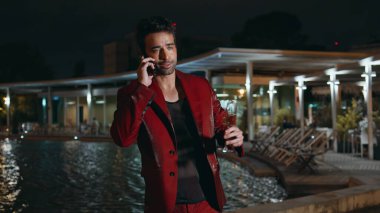 Havuz başı partisinde akıllı telefondan arayan göz kamaştırıcı adam kapat. Gülümseyen, maço, cep telefonuyla konuşan, dışarıda şampanya bardağı tutan. Şık kırmızı elbiseli mutlu iş adamı sohbetin tadını çıkar..