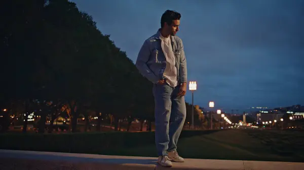 Stylish man relaxing at evening town park illuminated street lights. Attractive relaxed guy walking on concrete railings at windy night alone. Young hipster in jeans outfit enjoy weekend twilight.