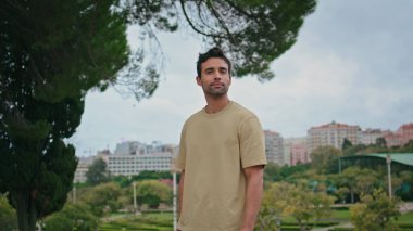 Latin adam yeşil parkı şehrin önünde poz veriyor. Yakından. Tıraşsız yakışıklı bir adam ağaç yeşilliğinin altında tek başına dikiliyor. Kasaba bahçesinde hafta sonu dinlenen kaygısız bir turist..