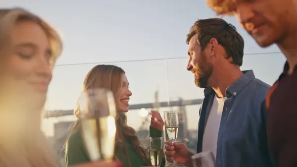 Smiling millennials drinking sparkling wine at rooftop party close up. Bearded cheerful man flirting with beautiful woman on sunny roof. Happy carefree friends holding champagne glass enjoying hangout