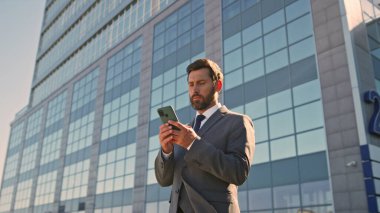 Odaklanmış patron ofis binasının yakınındaki cep telefonu ekranına bakıyor. Yakınlaştır. Zarif takım elbiseli adam açık havada akıllı telefondan iş e-postalarını okuyor. Ciddi iş adamı sokaklarda geziyor.