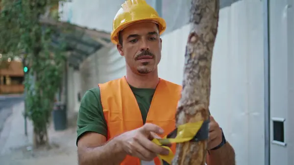 Focused guy putting warning tape at tree closeup. Confident builder man enclosing construction site with special stretch at greenery neighbourhood. Serious foreman in protective uniform working alone