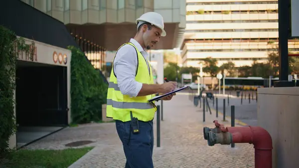 Building surveyor writing notes in folder working outdoors. Serious foreman in reflective vest uniform checking pipe standing at downtown. Professional man inspector examining house communications