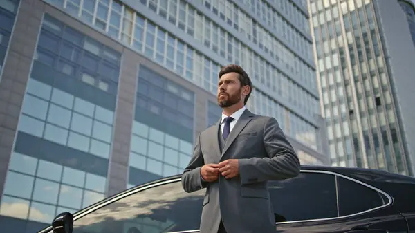 Millionaire posing at luxury car looking camera confidently zoom out. Elegant representative businessman standing at expensive auto near street skyscraper. Rich man buttoning jacket enjoying success.