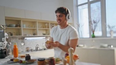 Modern apartman mutfağında kulaklıkla yemek pişiren komik bir adam. Neşeli, rahat adam kahvaltı için kadeh kaldırıyor kablosuz kulaklıkla müzik dinliyor. Mutlu yakışıklı, eğlenceli şarkı.