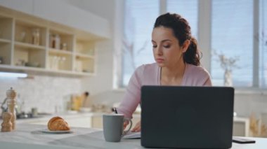 Modern dizüstü bilgisayarla rahat bir mutfakta oturan ciddi bir serbest çalışan kız. Ev ofisinde bilgisayar kullanarak uzaktan çalışan güzel bir iş kadını. Not defterine not alan, kahve içen odaklanmış bir kadın.