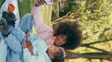 Afrikalı Amerikalı aile doğada akıllı telefonla video üzerinden konuşuyor. Mutlu, gülümseyen bir anne ve tatlı bir kızı telefon kamerasına dikey video konuşuyor. Kaygısız kızlar yeşil çimlerde güneşli çayırlarda oturuyor.