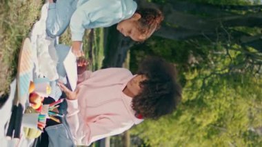 Aile piknik battaniyesinde güneşli yeşil doğada birlikte eğleniyor. Annesi dikey olarak vurulmuş, kağıda resim çizen sevimli Afrikalı Amerikalı çocuk. Rahat küçük kız çayır çimlerinde yaratıcılığın tadını çıkar..