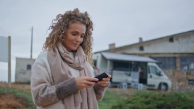 Tatilci kadın modern akıllı telefondan sonbahar fotoğrafı çekiyor. Mutlu kıvırcık kadın cep telefonuyla resim çekiyor. Rahat turist elinde cep telefonu karavanın yanında duruyor..