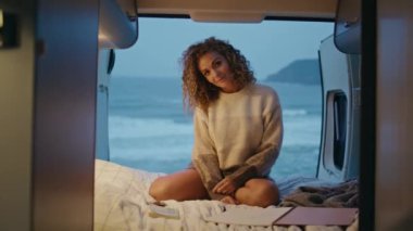 Akşam karavanı pozu veren kadın rahat bir yatakta oturuyor. Kaygısız hassas bayan kasvetli okyanus kıyısındaki karavanda dinlenen kameraya bakıyor. Romantik bayan turist, karavanda deniz tatilinin tadını çıkarıyor..