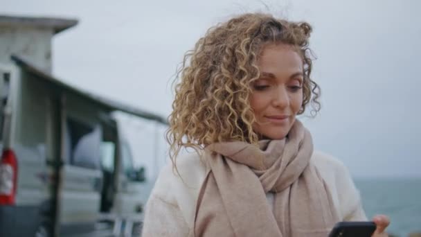 stock video Smiling lady typing sms on cellphone walking camper park at seashore gloomy day close up. Middle-aged curly woman looking smartphone screen at marine vacation. Happy tourist browsing phone on seacoast