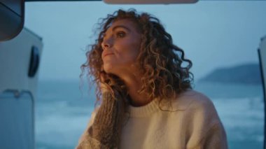 Romantik gezgin, akşam okyanus kıyısında, kapısı açık bir şekilde karavanda poz veriyor. Hayalperest kadın karavanda dinlenen kameraya bakıyor. Güzel bayan kampçıyla deniz yolculuğunun tadını çıkarın..