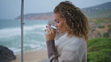 Ocean Beach 'te tatilde kupadan yudumlayan düşünceli bir kadın. Hayalperest kıvırcık kadın kahve içiyor seyahatte deniz güzeline bakıyor. Romantik kadınlar deniz kıyısında bulutlu deniz haftasonunun tadını çıkarırlar..