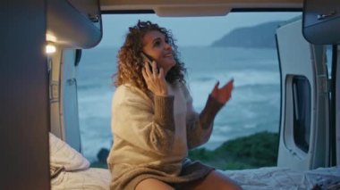 Mutlu kamp turistleri, kasvetli deniz kıyısındaki karavanda akıllı telefonları arıyor. Gülümseyen kıvırcık kadın cep telefonuyla konuşuyor deniz kıyısında rahat bir karavanda oturuyor. Olumlu bayan iletişimi sever..