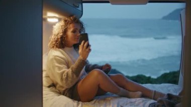 Kaygısız kadın Alacakaranlık Okyanusu sahilindeki karavanı arıyor. Gülümseyen kıvırcık kadın, akşam deniz kıyısında karavanda cep telefonuyla konuşuyor. Akıllı telefon konuşmalarıyla rahatlamış turistlerin haberleri