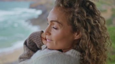 Güzel, olgun bir bayan bulutlu deniz kıyısında kameraya bakıyor. Haftasonu rüzgarlı okyanus kıyısında uzanan rahat kadın portresi. Çekici kadın sahilde tatilin tadını çıkarıyor.