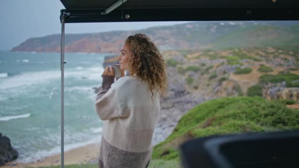 コーヒーカップ付きのトレーラーテラスに立っている海の波を見る幸せな観光客 ホットドリンクでマグカップを持っている笑顔の女性は曇った海岸を楽しみます キャンパーパークで海岸でリラックスしたキュリーな美しい女性 — ストック動画