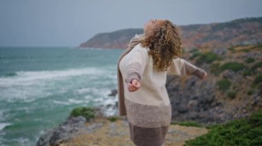 Deniz tatilini bulutlu kayalık kıyılarda geçiren bir kadın. Kaygısız çekici kadın taze deniz rüzgarının tadını çıkarırken el kaldırıyor. Kasvetli deniz kıyısında fırtınalı dalgalarda duran mutlu kadın gezgin..