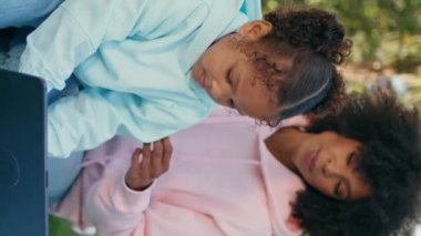 Afro-Amerikan ailesi yeşil parkta tabletle rahatlıyor. Mutlu, rahat bir anne, sekme oyunundaki dikey videoya odaklanmış şirin kızına bakıyor. Neşeli kızlar çayırda birlikte hafta sonunun tadını çıkarıyor.
