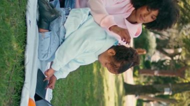 Anne, kızının piknik tabletinde oynamasını izliyor. Rahat bir Afrikalı Amerikalı kadın açık havada dikey çekime odaklanmış küçük bir kıza hayran. Güzel kıvırcık çocuk doğada klavye sekmesine basıyor.