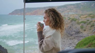 Deniz kıyısında çay içen bir bayan yakın mesafede mutlu deniz tatili yapıyor. Çekici kıvırcık kadın sonbahar haftasonu okyanus kıyısında kahvesini yudumluyor. Gülümseyen kadın turist, kameraya bak özgürlük hisset.