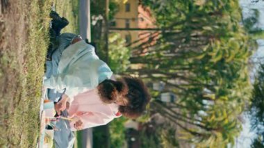 Yeşil Park 'ta Afro-Amerikan aile pikniği. Rahatlamış genç bir anne, güneşli çayır çimlerinin üzerinde oturan küçük bir kızla dikey videoda eğleniyor. Kıvırcık kadın, sevimli kızıyla hafta sonunun tadını çıkar..