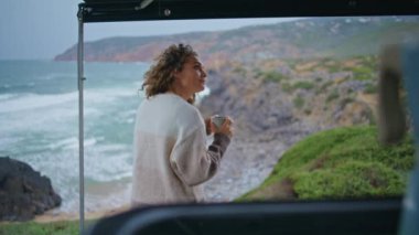 Kaygısız kadın, karavanın yanında duran deniz dalgalarından hoşlanır. Gevşemiş kıvırcık kadın kıyıda kahve içiyor bulutlu deniz manzaralı. Mutlu bayan gezgin karavan parkında deniz tatili yapıyor..