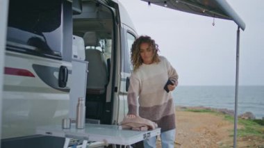 Sakin gezgin kamp yerinde rahat bir eşarp eşarbıyla sarıp sarmalayarak sonbahar günü. Deniz kampı tatilinde kasvetli deniz manzarasına bakan rahat olgun bir kadın. Akıllı telefon tutan kıvırcık kadın karavandan çıkıyor..