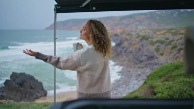 Deniz kıyısında elinde kahve fincanı tutan romantik bir bayan. Kıvırcık, rahat bir kadın okyanus kıyısında karavanın yanında sıcak çay içiyor. Çekici bayan turist yağmuru kontrol etmek için uzanıyor. Deniz yolculuğu konsepti.