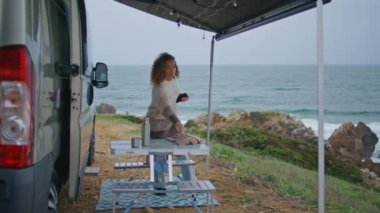 Güzel okyanus kıyısına giden bir kadın. Eşarp takan kıvırcık, gevşemiş kadın bulutlu deniz kıyısında kamp tatilinin tadını çıkarıyor. Kaygısız kadın turist karavan terasında dinleniyor..