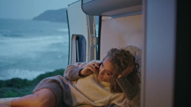 夜の海岸で寝台のモーターホームに横たわっている女性観光客が近づく トレーラーでリラックスして電話するケアフリーカーリーレディ 笑顔の女性旅行者はキャンプ休暇で会話を楽しむ — ストック動画