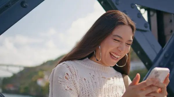 Laughing model reading sms on bridge closeup. Attractive asian woman holding modern smartphone watching funny videos outdoors. Stylish brunette girl typing mobile phone standing near iron construction