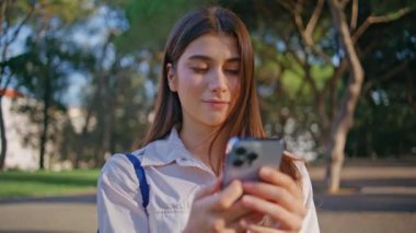 Gülümseyen öğrenci cep telefonuna göz atıyor. Parka yakın çekimde teknoloji tarafından özümseniyor. Neşeli genç bir kadın akıllı telefondan sosyal medyayı izliyor. Güneşli doğada duruyor. Telefon mesajı okuyan mutlu kız..