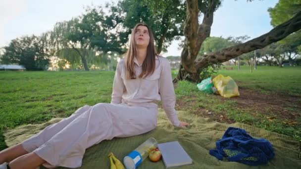 心神不宁的女人在清理完生态行动后 喜欢在公园野餐 女孩躺在被绿色大自然环绕的毯子上放松 背负着环保的垃圾袋 有思想的年轻女士躺在草地上 — 图库视频影像