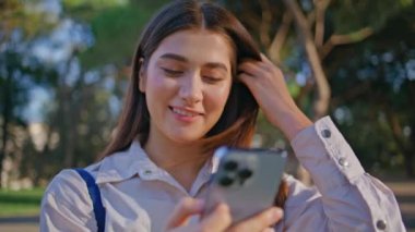 Gülümseyen iş kadını canlı yeşil parkta akıllı telefonuna göz atıyor. Güzel bir yaz parkında cep telefonuyla mesajlaşan mutlu kadın. Teknolojinin zevkini vurgulayan güneşli arkaplan. 