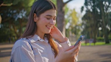 Güneşli parkta cep telefonuna mesaj atan neşeli kadın dijital çağda mutluluğu resmediyor. Portre hanımı dışarıda teknolojiyle eğleniyor. Gülümseyen kız yeşil ağaçların altında akıllı telefondan mesaj atıyor.