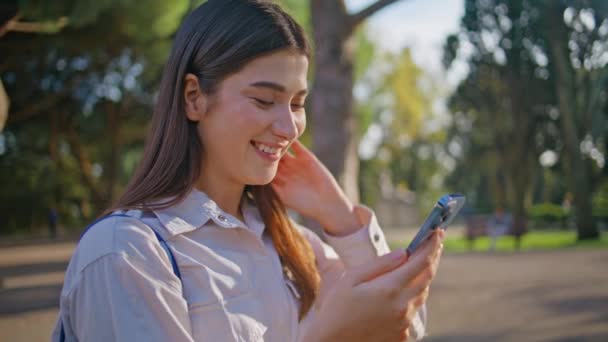 陽気な公園で陽気な女性メッセージング携帯電話は デジタル時代の幸せを描いています アウトドアテクノロジーでレジャーを楽しむポートレートレディ 緑の木の下でスマートフォンをテキストする笑顔の少女 — ストック動画