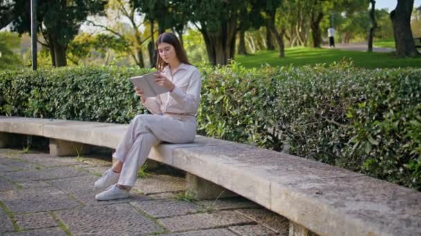 平和を伝える日光公園の本を読んでいる少女 緑の自然の中でベンチに座っている興味深い文学と余暇の時間を楽しむ若い個々の女性 緑でリンゴを食べる女性リーダー — ストック動画
