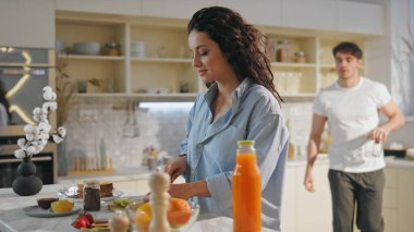 Genç çift kahvaltıyı evde hazırlıyor. Yakın çekim. Güzel bir kadın sıcak mutfakta taze elma dilimliyor. Gülümseyen yakışıklı erkek arkadaş portakal suyu için gözlük alıyor. Mutlu evlilik kavramı. 