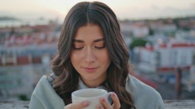 Günbatımı şehir manzaralı balkon resminde espresso içen nazik bir kadın. Sakin bir model, rahat otel manzaralı terasta içecek görünümlü kamera çekiyor. Gülümseyen şehvetli kız rahatlıyor akşamın tadını çıkarıyor. 