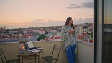Romantik kız güneşin doğduğu şehrin balkonunda kahve içiyor. Sabah terasında elinde bardakla duran mutlu bayan. Gülümseyen kadın kasabayı düşünüyor yalnız başına dinleniyor. Esmer model çayını yudumluyor 