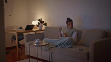 Akşamları video oyunu oynayan odaklanmış bir kadın koltukta oturuyor. Duygusal kız elinde joystickle cep telefonu mesajlarını kontrol ediyor. Oyun tahtası kullanan pozitif bir kadın. Oturma odasında kanepede eğleniyor. 