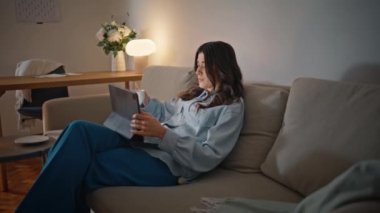 Akşam kızı rahat ev çekiminde Pad Screen seyrediyor. Rahat kadın tablet videosu izliyor ve sakince dinleniyor. Esmer kadın çayını yudumluyor. Lambanın içindeki rahat koltukta oturuyor. Ev içi rutin