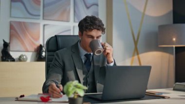 Yansıtıcı iş adamı modern ofis ortamında kahvesini yudumluyor. Düşünceli profesyonel dinlenme anının tadını çıkarıyor masada sıcak içecek içiyor. Düşünceli resmi adam tek başına notlar yazıyor.
