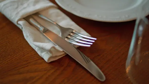 Elegant cutlery lying on table cafeteria ready for use close up. Metal fork knife neatly wrapped in white napkin placed on wooden desk cozy restaurant. Empty served place for dinner. Tableware concept