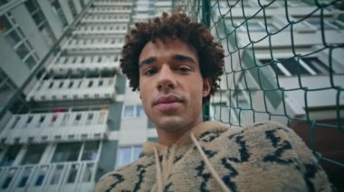 Yüksek apartmandaki kameraya bakan Latin Amerikalı adamın portresi. Yakışıklı kıvırcık adam şık bir kapüşonluyla sokakta poz veriyor. Gülümseyen gevşetici hipster tasasız şehir haftasonunun tadını çıkar..