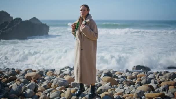 居心地の良いコートで岸に立っている若い女性 海の波が近くの岩の海岸に衝突する 海岸線で静かな熟考でポーズする貧しい探検家 ファッショナブルなモデルは 屋外で平和な自然を楽しむ — ストック動画