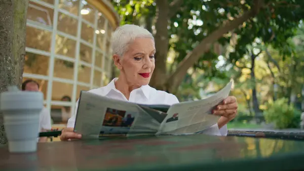 Mature lady looking magazine enjoying summer morning in open air cafe closeup. Beautiful elderly businesswoman reading press sit restaurant table. Successful senior with red lips rest weekend alone.
