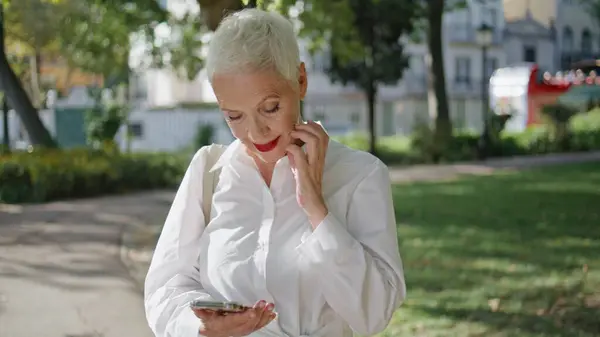 成熟的女士在阳光充足的公园特写镜头中键入浏览手机 微笑的退休人员在户外看手机短信 时尚的老年女商人在夏日花园用智能手机在线发短信 免版税图库图片